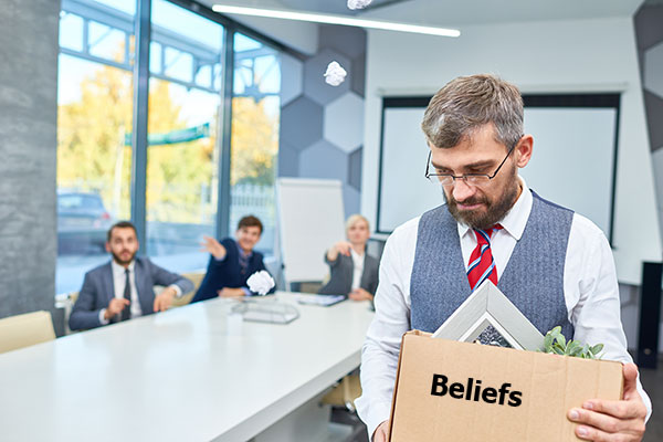 man leaving an office with a box of stuff. On the box, it says beliefs. Co-workers in the background are pointing and laughing